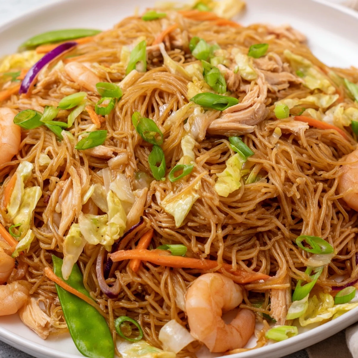 Family-style serving platter of Traditional Pancit Bihon garnished with fresh scallions and lemon wedges alongside meat and vegetables